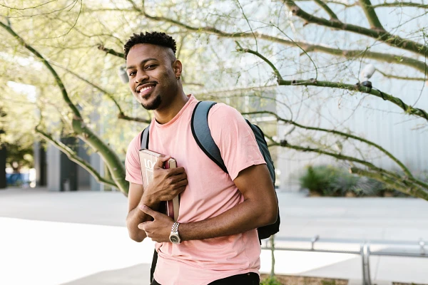 College student wearing a backpack and pink t-shirt, holding a textbook, standing outdoors on college campus